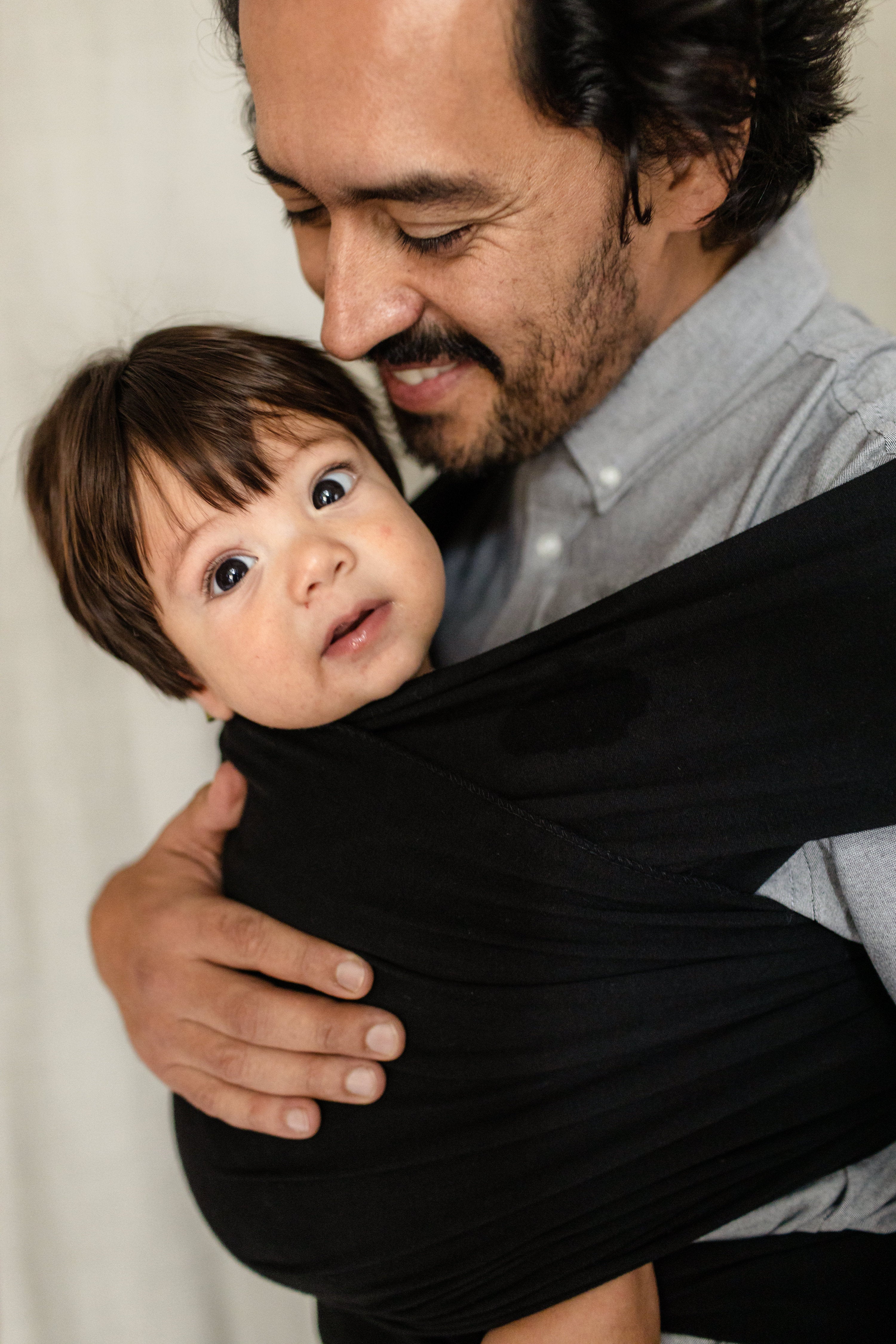 Happy dad is proudly looking at his son's smiling face while he wears him in the black boba classic wrap in the hip healthy ergonomic front carry position. 