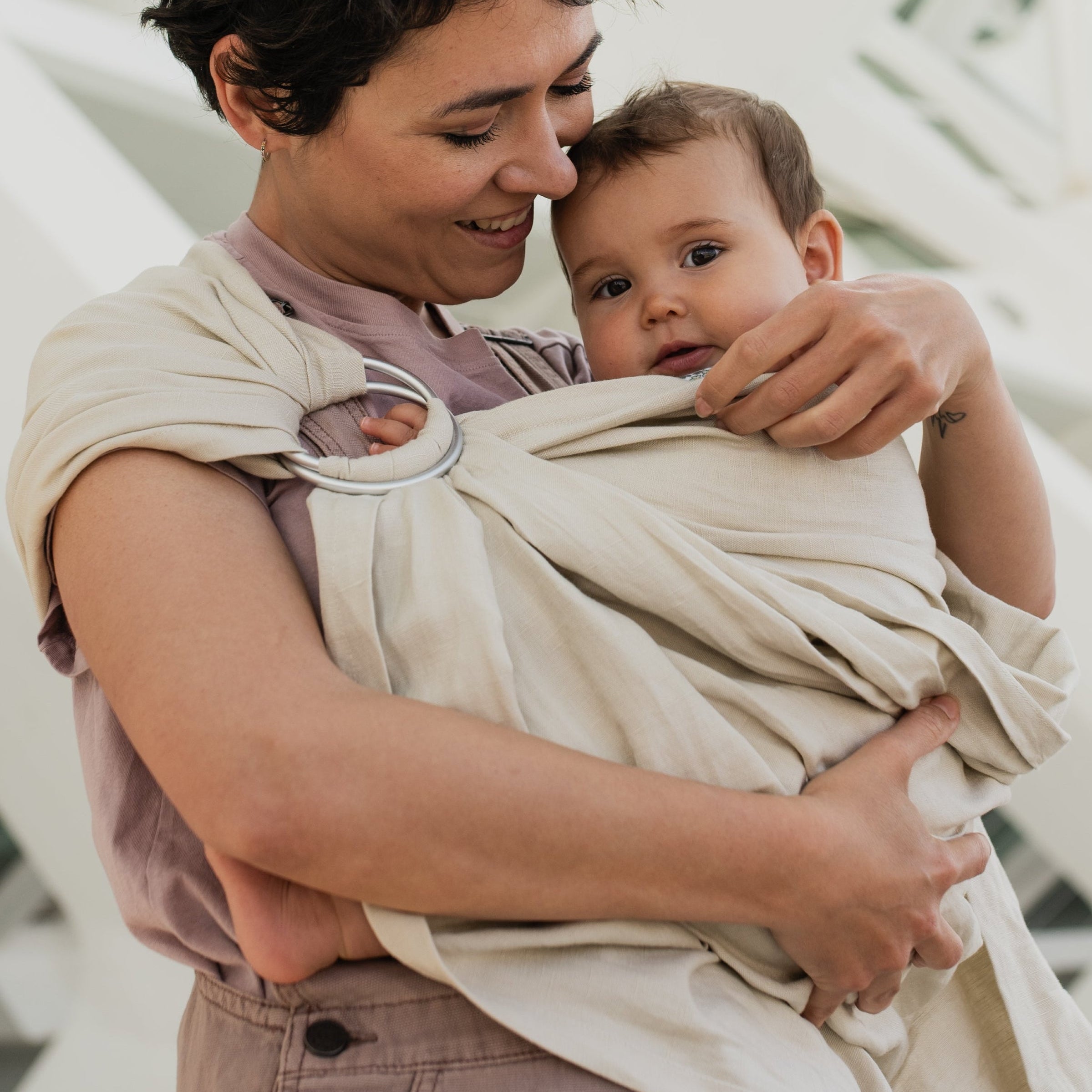 A woman wearing a beige baby carrier wrap holds a baby in a neutral-toned setting in front of a modern white geometric building.