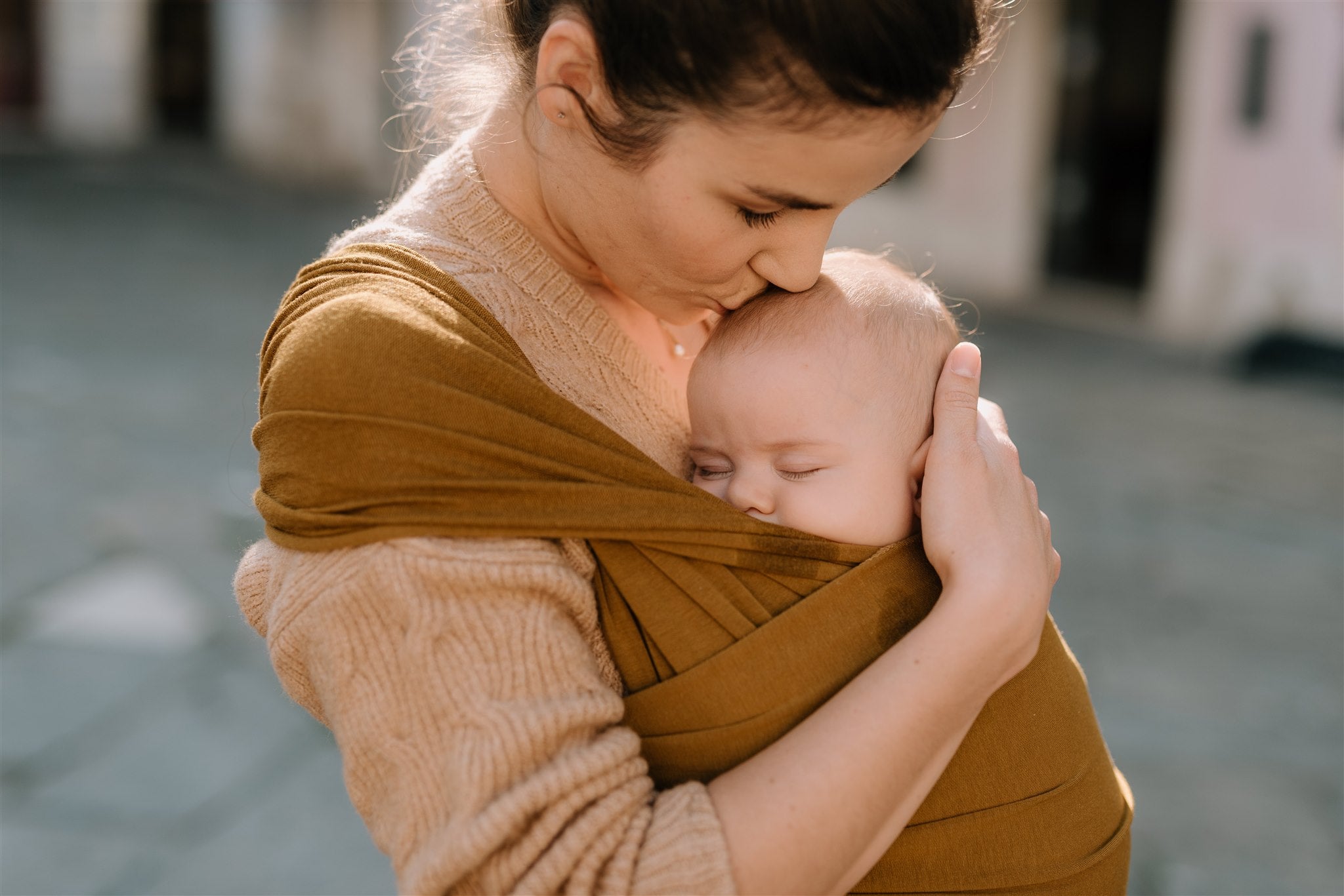 A happy loving mom is kissing her sleeping daughter's head while she takes a nap in the mustard yellow serenity wrap bamboo.
