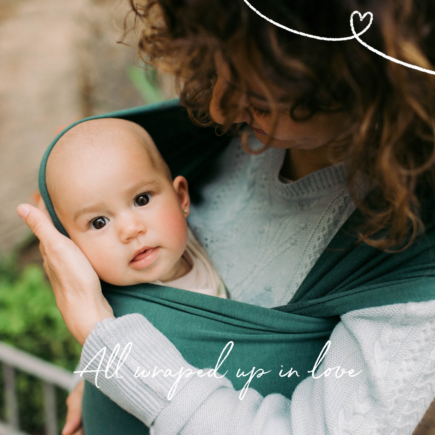 A confident smiling brown curly haired mom is holding her cute baby girl in a green Rainforest bamboo Serenity Wrap. 