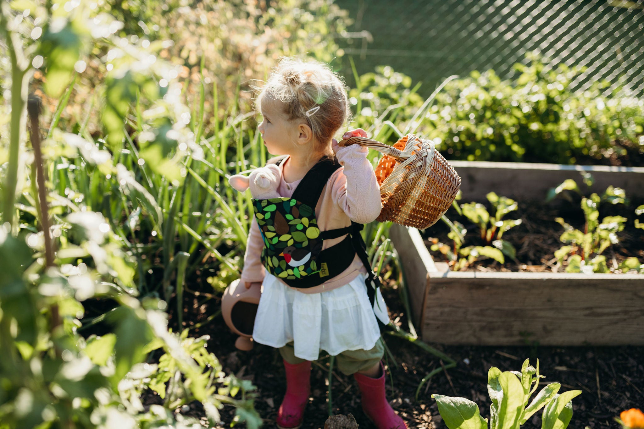 A young blonde girl with two ponytails standing in the garden holding a basket in her hand and wearing her stuffed toy pig in her Boba Mini carrier. 
