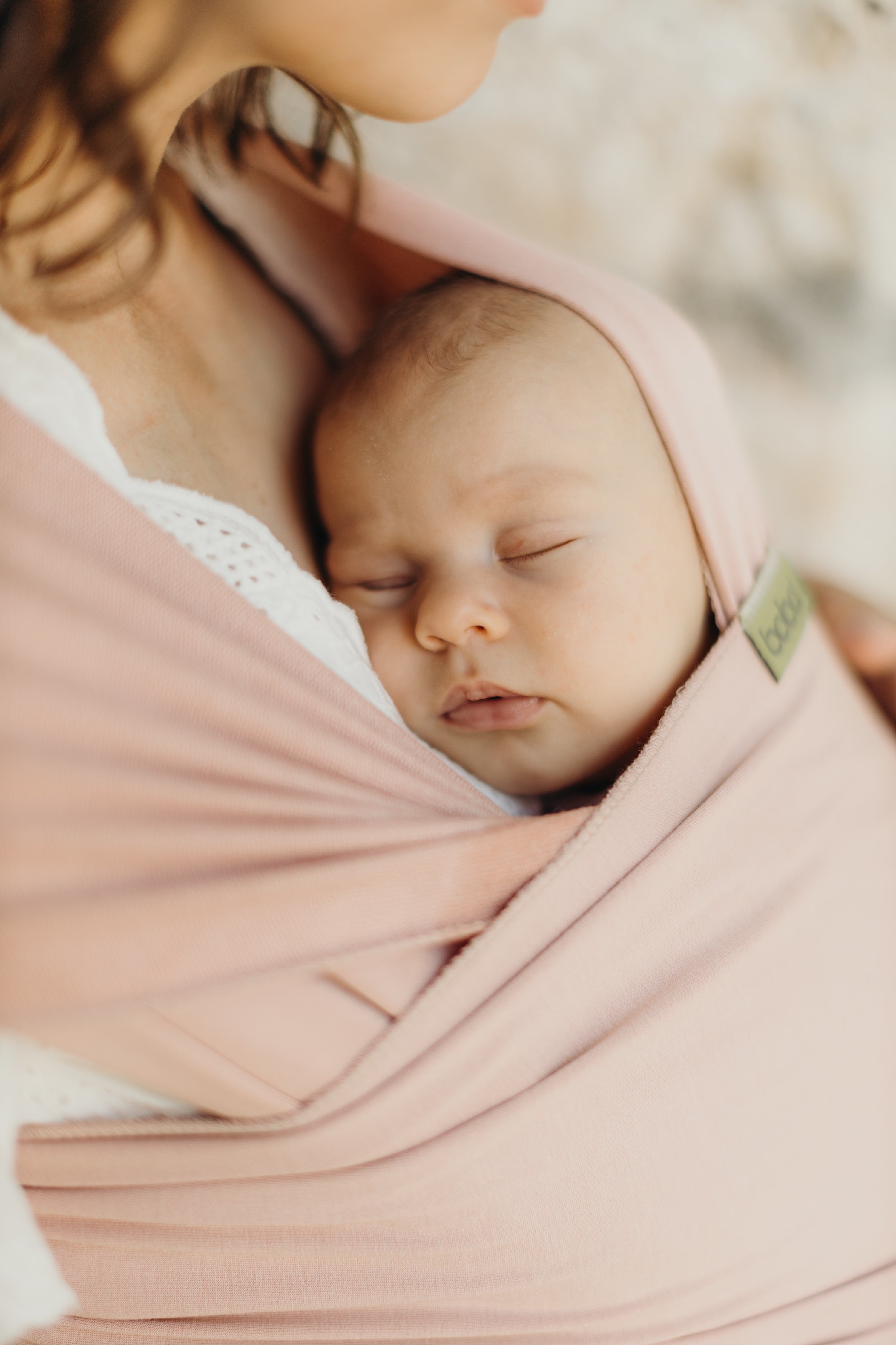 Cute sleeping baby girl is resting her tiny head on her mother's chest as she is wearing her in the boba wrap serenity bloom in an ergonomic position.